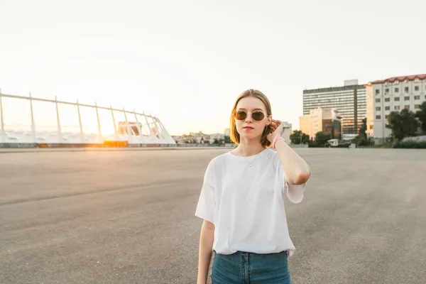 Ritratto ragazza elegante in occhiali e vestiti alla moda posa sullo sfondo di un paesaggio urbano al tramonto, guardando in una fotocamera con un volto serio. Bella ragazza di strada in posa — Foto Stock