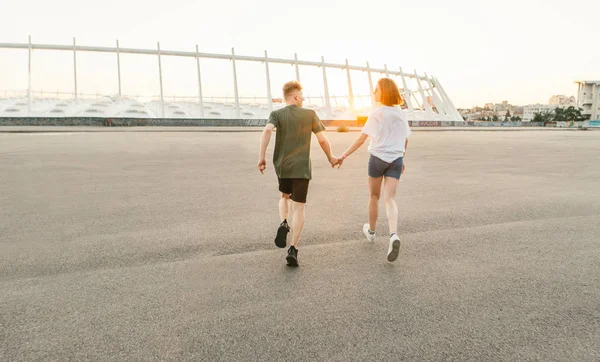 Bellissima coppia in abiti alla moda gestita tenendosi per mano sullo sfondo del tramonto e del paesaggio urbano. Foto in stile leggero. Coppia amorevole correre, guardare dal retro . — Foto Stock