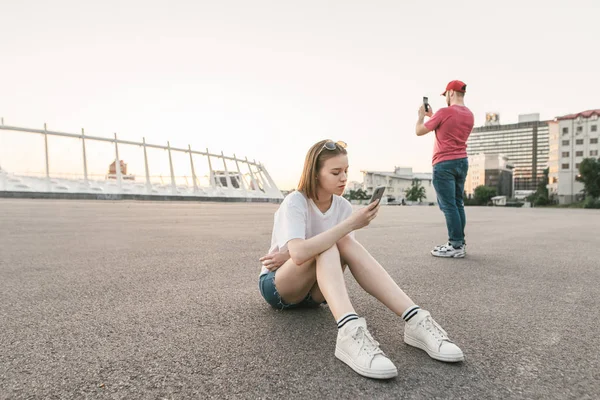 在城市风景的背景下,人们在户外使用智能手机。女孩使用智能手机,一个男人做照片。千禧一代手里拿着小玩意儿散步. — 图库照片