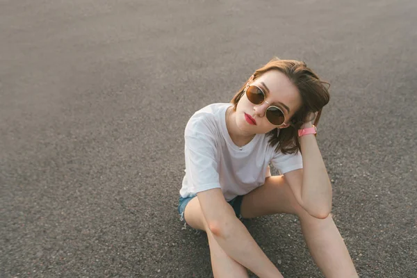 Şık bir rahat giyim ve güneş gözlüğü seksi bir kız Trendy sokak portresi sokakta oturur ve kamera bakar, asfalt arka planda. Bir kızın akşam yaşam tarzı portresi. — Stok fotoğraf