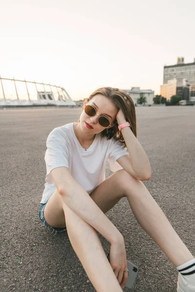 Menina elegante em óculos e uma camiseta branca se senta no pavimento e olha para a câmera com um rosto sério. Retrato de rua de uma menina sexy no fundo de uma paisagem da cidade à noite . — Fotografia de Stock