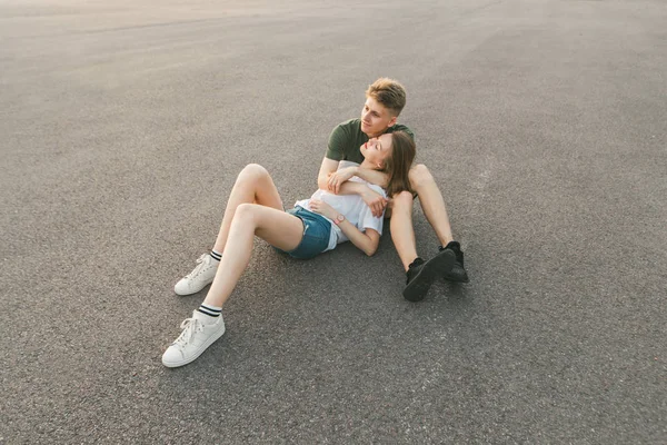 Foto de jovem casal feliz sentado no asfalto, abraçando e olhando para longe. Bonito casal de rua roupas elegantes sentado no fundo da capa do asfalto, vista de cima — Fotografia de Stock