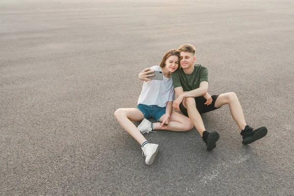 Komik çift kaldırımda oturan ve bir akıllı telefon üzerinde sephi yapıyor seviyor. Şık genç adam ve kız sokakta oturup akıllı telefonda bir fotoğraf yapmak, komik yüzleri ile kameraya poz.