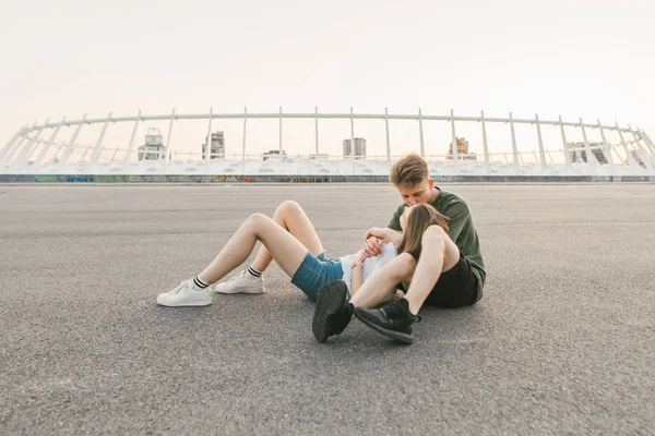 Portret van een mooi paar ontspannen in de straat op de achtergrond van de moderne architectuur, zittend op de stoep in een eenzame plaats en knuffelen, het dragen van een stijlvolle casual kleding. Liefdesverhaal. — Stockfoto