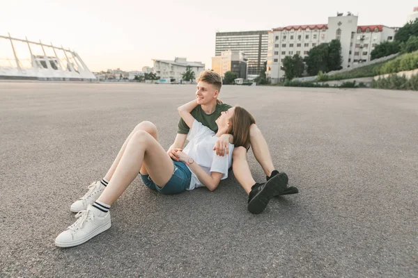 Gelukkige gelukkige paar ontspannen terwijl zittend op de grond, de jonge man knuffels een meisje, ze lachen en kijken opzij, het dragen van stijlvolle Streetwear. Een prachtig paar knuffelen in een wildernis buiten de stad. — Stockfoto