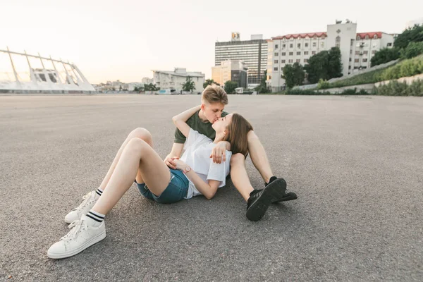 Retrato de um casal bonito e elegante. Bonito casal engraçado abraçando e beijando, menina deitada de joelhos no cara, jovem sentado no asfalto no fundo da paisagem da cidade . — Fotografia de Stock