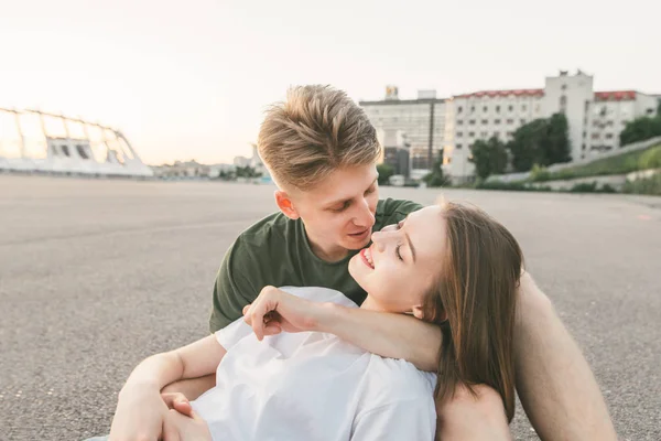 Foto in primo piano di una giovane coppia che abbraccia e sorride sullo sfondo della città. Coppia amorevole seduta su asfalto, paesaggio cittadino e parlando, ragazzo sorridente abbracciare una ragazza felice. Ritratto stradale — Foto Stock