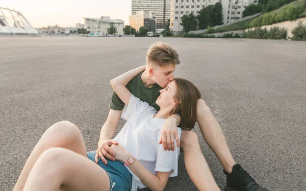 Liefdevolle paar knuffels en kusjes liggen op de stoep tegen de achtergrond van het stadslandschap. Jonge man kussen een vriendin in het voorhoofd, het meisje glimlacht. Liefdesverhaal. Straat foto. — Stockfoto