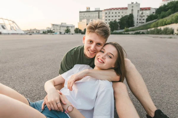 Bella coppia felice seduta sul marciapiede, sullo sfondo del paesaggio della città, abbracciando e guardando in camera.Street ritratto di una giovane coppia sorridente in abiti eleganti poggiata sulla strada — Foto Stock