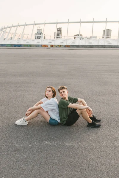 Mooi stijlvol paar zittend op het dak naast elkaar, kijkend naar de camera en glimlachend. Jonge man en meisje rusten zittend op asfalt. Lifestyle foto van een mooi paar. Verticale foto. — Stockfoto