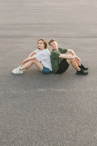 Vrolijk paar zittend naast elkaar op het asfalt, kijkend naar elkaar en glimlachend. Mooi paar rusten terwijl zittend op de grond. — Stockfoto