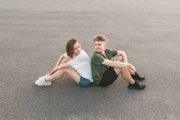 Ein fröhliches Paar, das nebeneinander auf dem Asphalt sitzt, einander ansieht und lächelt. schönes Paar ruht sich auf dem Boden aus. — Stockfoto