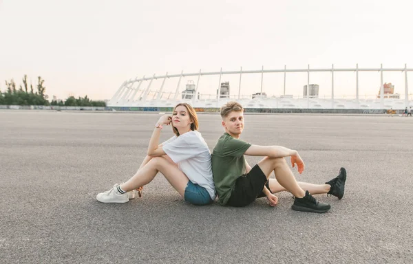 Mooi stijlvol paar zittend op het dak naast elkaar, kijkend naar de camera en glimlachend. Jonge man en meisje rusten zittend op asfalt. Lifestyle foto van een mooi paar. — Stockfoto