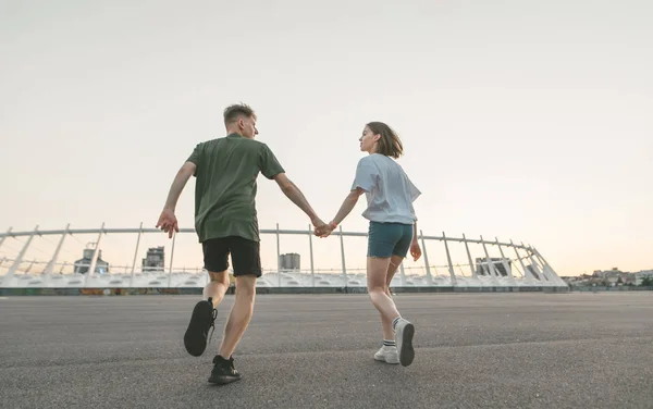 気楽な二人の若者は、夕方の街の風景を背景に、手をつないで、お互いを見つめ、後ろから微笑みながら走った。愛するカップルのライフスタイル写真ランニング — ストック写真