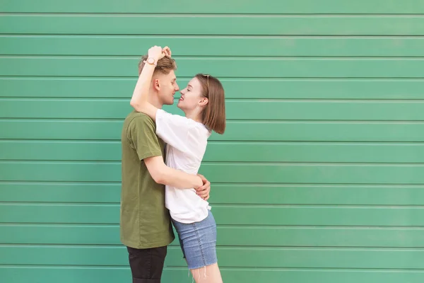 Mooie jonge paar kerel en meisje knuffelen op groene achtergrond, kijkend naar elkaar en glimlachend. Straat portret van een mooi jong stel in een casual jurk tegen een achtergrond van een groene muur. — Stockfoto