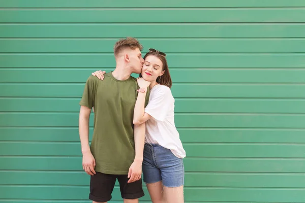 Фото влюбленных пар в повседневной одежде на фоне зеленой стены, молодой человек целует девушку, она улыбается и довольствуется. Любящая девушка и парень на цветном фоне . — стоковое фото