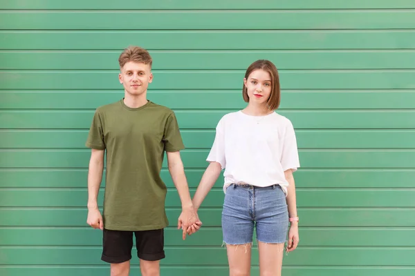 Um jovem e uma garota atraente dão as mãos em um fundo azul-turquesa e olham para a câmera. Bonito jovem casal de pé no fundo de uma parede verde e posando na câmera — Fotografia de Stock