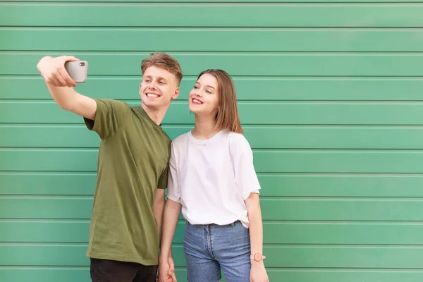 Un cuplu fericit tânăr și o fată fac selfie pe fundal turcoaz, uitându-se la cameră și zâmbind smartphone-ul. Omul face selfie cu prietena lui pe fundalul unui perete verde . — Fotografie, imagine de stoc