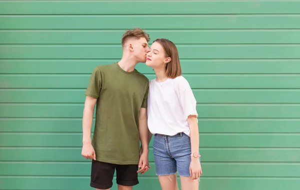 Bela jovem casal em um vestido casual elegante em pé sobre um fundo verde, um menino beija uma menina em sua bochecha, ela está satisfeita.Jovem beija uma menina em um fundo turquesa, conceito de amor — Fotografia de Stock