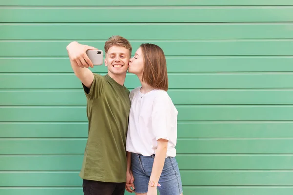 Любящая молодая пара делает селфи на цветном фоне, девушка целует мальчика, молодой человек улыбается и смотрит в камеру смартфона, носить стильную повседневную одежду. Любовь - это понятие . — стоковое фото