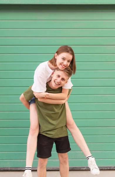 Leende ung man bär casual kläder bär en lycklig flicka på ryggen, tittar på kameran och har kul. Snygg par ha kul på bakgrunden av en turkos vägg. Vertikalt foto. — Stockfoto