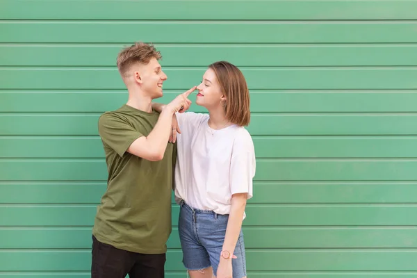 Street photo of a stylish young couple standing on the background of a green wall,young man touches the girl\'s nose and smiles.Happy couple, street photo,isolated on the background of a turquoise wall