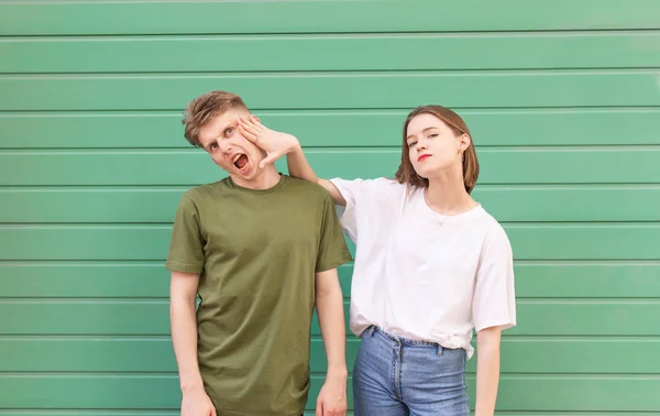 Engraçado jovem casal em um fundo verde colorido, a menina segura o cara por trás de seu rosto, ele não gosta. Casal alegre, elegante, retrato de rua em um fundo turquesa . — Fotografia de Stock