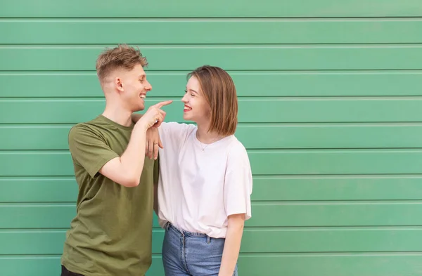Vackra unga paret kille och flicka på grön bakgrund, titta på varandra och leende. Gatuporträtt av ett vackert ungt par i ett fritidskläder mot en bakgrund av en turkos vägg — Stockfoto