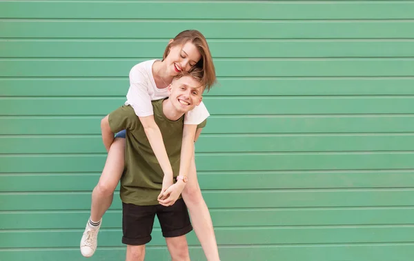 Sonriente joven con ropa casual lleva una chica feliz en la espalda, mira a la cámara y se divierte. Pareja con estilo que se divierten en el fondo de una pared de color turquesa. Copiar espacio —  Fotos de Stock