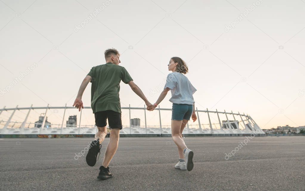 Carefree couple of young people ran holding hands, looking at each other and smiling, looking from the back,against the backdrop of an evening city landscape.Lifestyle photo of a loving couple running