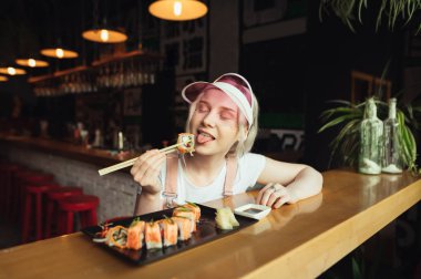 Sevimli pembe giysiler içinde Çekici kız bir Japon restoranında oturur ve yemek çubukları ile suşi yiyor, gözleri kapalı memnun olur. Bir Japon restoranında suşi rulo yiyen sevimli kadın.
