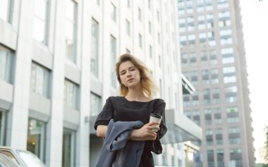 Çekici genç kadın şehirde dolaşan bir fincan kahve tutarak kameraya bakıyor. Sokaktaofisyon binasının önünde poz veren güzel bir kızın portresi. Kentsel kavram.