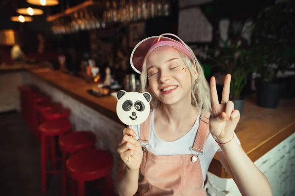 Retrato de uma menina bonito dentro de casa vestindo uma roupa rosa, maquiagem com brilho em seu rosto, segurando um pirulito panda na mão, mostrando sinal de paz com sua mão.Menina cosplayer posando na câmera. Estilo coreano — Fotografia de Stock