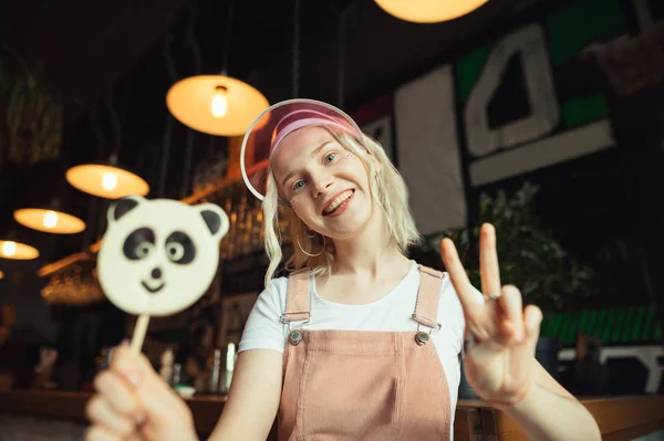 Portrét šťastné dívky v růžových roztomilým oblečení, které drží Panda Lollipop, dívá se do kamery, usmívá se a pózuje mírovým znamením. Dívka s cukrovinkou v ruce. — Stock fotografie