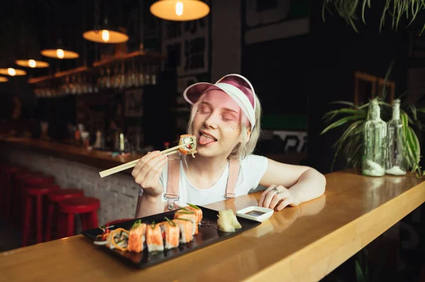 Atraktivní dívka v roztomilým růžovém oblečení sedí v japonské restauraci a sní sushi s hůlky, je spokojená se zavřenýma očima. Roztomilá žena, která žere sushi v japonské restauraci. — Stock fotografie