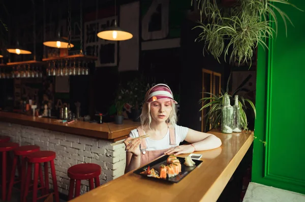 Jolie adolescente assise à une table dans un restaurant asiatique, mangeant des rouleaux de sushi sur des étagères, concentrée et savoureuse nourriture japonaise. Dame manger des rouleaux de sushi dans un restaurant japonais . — Photo