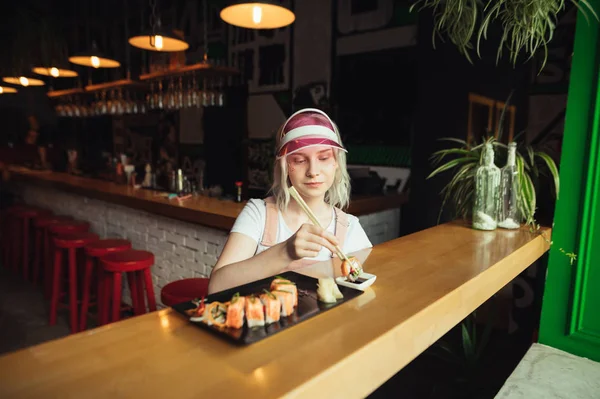 분홍색 옷을 입은 귀여운 소녀와 모자는 레스토랑에서 초밥을 먹고, 그녀는 일본 요리를 좋아합니다. 아름다운 소녀는 초밥롤과 함께 막대기를 들고 아시아 레스토랑에 앉아 간장 sauc에 담그고 — 스톡 사진
