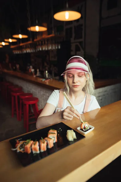 Menina bonita senta-se em um restaurante asiático segurando paus com um rolo de sushi e mergulhando-o em molho de soja. Menina bonito em roupas cor de rosa comendo um conjunto de sushi em um restaurante, ela adora cozinha japonesa — Fotografia de Stock