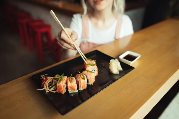 Фоновому режимі. Жінка їсть Апетитний суші рол в японському ресторані, бере роли з паличками, обрізаний фото зверху, дівчина руками приймають суші Рулетики з паличками з суші плита. — стокове фото