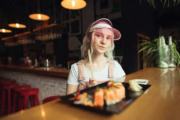 Retrato de una hermosa chica sentada en una mesa en un restaurante con un plato de rollos de sushi, sosteniendo palillos en sus manos, mirando a la cámara con una cara seria. Linda dama cena para sushi — Foto de Stock