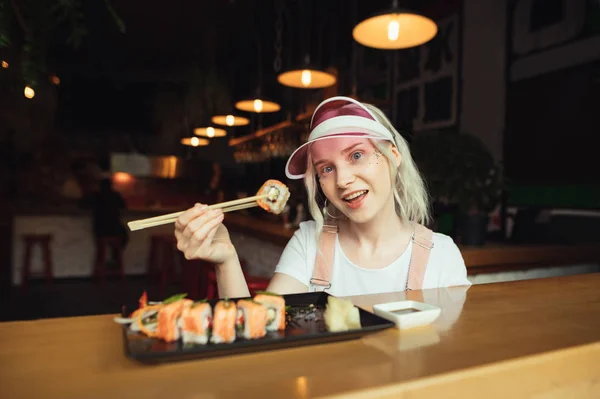 Portret uśmiechniętej dziewczyny z pałeczkami i Sushi Roll, siedząc w restauracji, jedzenie kuchni japońskiej, patrząc na aparat i stwarzające. Atrakcyjna dama w różowej czapce pokazuje Sushi Roll na pałeczki w aparacie — Zdjęcie stockowe