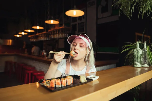 Linda chica con maquillaje y gorra rosa come sushi con palillos en un acogedor restaurante asiático. La señora muerde sushi con placer, disfruta comiendo sushi los fines de semana. Concepto de cocina japonesa . — Foto de Stock
