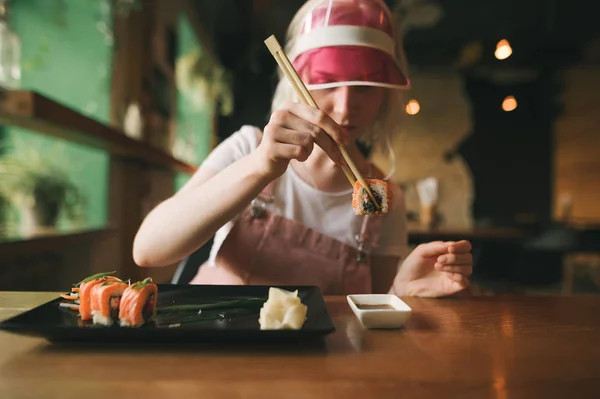 Девушка держит палочки с суши, макает суши-ролл в соевом соусе. Привлекательная женщина в розовой кепке ест суши-роллы палочками для еды. Близко. Фон . — стоковое фото