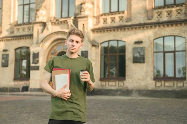 Портрет серьезного кавказского студента в зеленой футболке, стоящей перед университетом с копировальными книгами и кофе. Сконцентрированный спокойный студент, стоящий во дворе колледжа с кофе и книгами . — стоковое фото