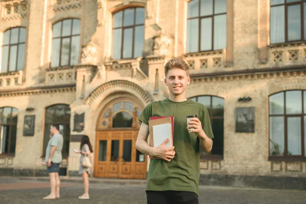 Красива молода студентка-чоловік стоїть перед університетською будівлею з блокнотами і забирає чашку кави. Усміхнений привабливий студентський хлопчик біля кампусу коледжу тримає книги та каву . — стокове фото
