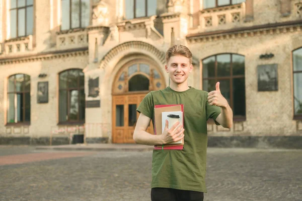 Молодой жизнерадостный успешный кавказский ботаник-студент показывает большой палец вверх, стоя с книгами, университет на заднем плане, рядом с кампусом, солнечный. Позитивный улыбающийся студент показывает пальцем вверх рядом с колледжем . — стоковое фото