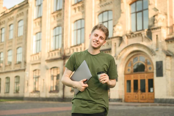 Улыбающийся молодой студент с ноутбуком смотрит в камеру на фоне университета. Уверенный молодой улыбающийся студент на открытом воздухе возле кампуса колледжа с кофе и блокнотом . — стоковое фото