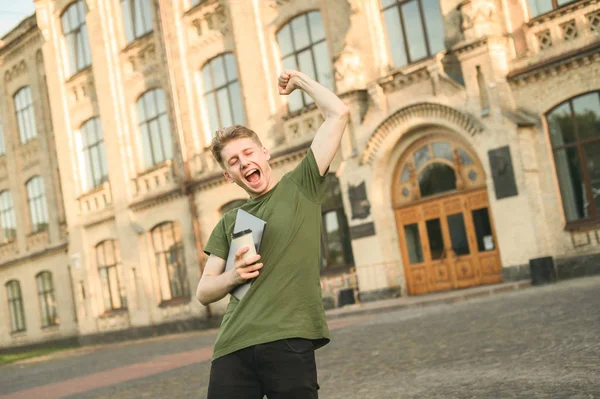 Boldog vidám izgatott férfi hallgató ünnepli győzelme a felvetett kezek előtt főiskolai campus. A fő feladat befejeződött, vizsgák alatt, elérte a sikert, győztes hallgató laptop és kávé. — Stock Fotó