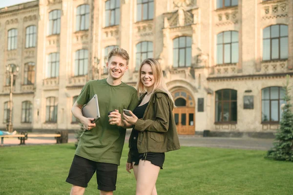 Casal de estudantes alegres andando depois de estudos, segurando laptop e smartphone, sorrindo, curtindo, rindo, ajudando uns aos outros com estudos. Todos vestidos com conforto casual desgaste, dia ensolarado . — Fotografia de Stock