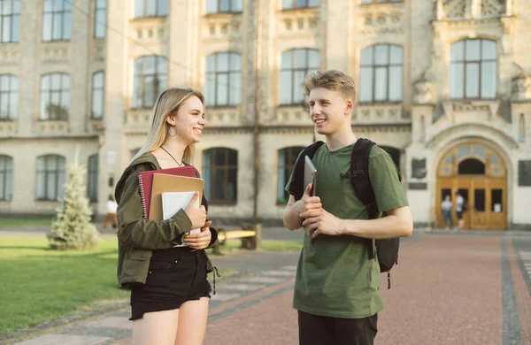 Casal alegre de estudantes universitários tendo uma pequena conversa perto do campus segurando livros, cadernos e laptop. Dois estudantes bonitos atraentes agradavelmente falando perto do prédio da universidade . — Fotografia de Stock